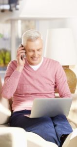 Elderly man working on computer with cell phone to his ear, smiling.